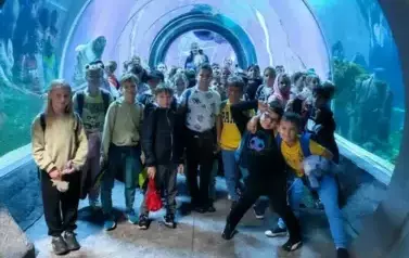 Grupa dzieci pozująca do zdjęcia na tle błękitnego oceanarium pełnego egzotycznych ryb.