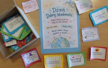 Na drewnianym stole znajduje się pudełko z kolorowymi, własnoręcznie wykonanymi karteczkami z dobrymi słowami. Obok leży plakat &bdquo;Dzień Dobrej Wiadomości&rdquo; z hasłem &bdquo;Dobre słowa są po to, żeby się nimi dzielić&rdquo;.