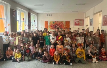 Grupa dzieci ubranych w jesienne stroje i ozdoby pozuje wsp&oacute;lnie na szkolnym korytarzu. W tle widoczne są jesienne dekoracje, kolorowe liście oraz prace plastyczne