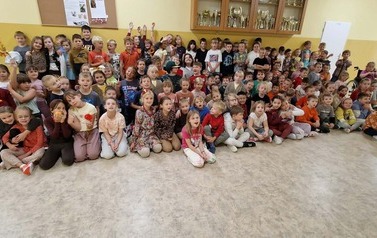 Duża grupa dzieci w jesiennych strojach i kolorach pozuje wsp&oacute;lnie na szkolnym korytarzu. Dzieci siedzą i stoją w kilku rzędach, a w tle znajdują się szkolne dekoracje i gabloty.
