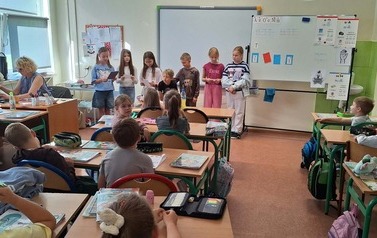 Uczniowie siedzą w klasie i słuchają kolegi czytającego książkę. Dzieci zgromadzone są w części sali, a na p&oacute;łkach w tle znajdują się książki i materiały edukacyjne.