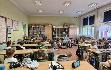 Uczniowie klasy trzeciej czytają młodszym kolegom w klasie. Kilkoro dzieci stoi przed tablicą z książkami w dłoniach, a pozostali uczniowie siedzą przy ławkach i słuchają.