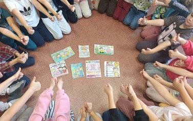Uczniowie siedzą w kręgu na podłodze i pokazują kciuki uniesione w g&oacute;rę. W centrum kręgu znajdują się kolorowe materiały dotyczące przeciwdziałania przemocy r&oacute;wieśniczej, między innymi plakaty z hasłami zachęcającymi do re