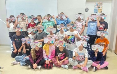 Grupa uczni&oacute;w siedzi i stoi w sali, prezentując wykonane przez siebie kolorowe papierowe okulary. Na okularach znajdują się wzory, napisy i ilustracje związane z tematyką Tygodnia Przeciwdziałania Przemocy R&oacute;wieśniczej.