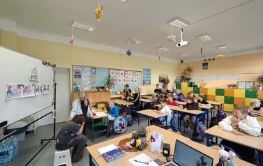 Uczniowie uczestniczą w zajęciach w sali lekcyjnej. Na tablicy widoczne są materiały dotyczące bezpieczeństwa.