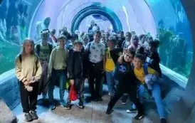 Grupa dzieci pozująca do zdjęcia na tle błękitnego oceanarium pełnego egzotycznych ryb.