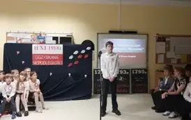 uczeń klasy sz&oacute;stej recytuje wiersz a z jego prawej i lewej strony siedzą uczniowie klas młodszych i starszych biorący udział w wieczornicy