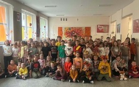 Grupa dzieci ubranych w jesienne stroje i ozdoby pozuje wsp&oacute;lnie na szkolnym korytarzu. W tle widoczne są jesienne dekoracje, kolorowe liście oraz prace plastyczne