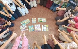 Uczniowie siedzą w kręgu na podłodze i pokazują kciuki uniesione w g&oacute;rę. W centrum kręgu znajdują się kolorowe materiały dotyczące przeciwdziałania przemocy r&oacute;wieśniczej, między innymi plakaty z hasłami zachęcającymi do re