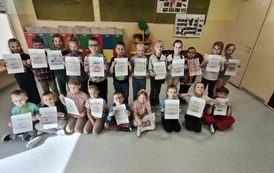Grupa uczni&oacute;w stoi w dw&oacute;ch rzędach w sali lekcyjnej i prezentuje wykonane przez siebie kolorowe prace. Dzieci trzymają kartki z ilustracjami i hasłami promującymi zgodę, przyjaźń oraz dobre relacje.