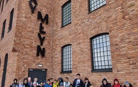 Uczniowie przed budynkiem Starego Młyna w Zgierzu. Przed wysokim, ceglanym budynkiem z dużymi oknami zabezpieczonymi czarnymi kratami pozuje grupa uczni&oacute;w klas 3c i 3d. Dzieci stoją i klęczą w kilku rzędach, ubrane w kolor
