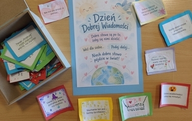 Na drewnianym stole znajduje się pudełko z kolorowymi, własnoręcznie wykonanymi karteczkami z dobrymi słowami. Obok leży plakat &bdquo;Dzień Dobrej Wiadomości&rdquo; z hasłem &bdquo;Dobre słowa są po to, żeby się nimi dzielić&rdquo;.