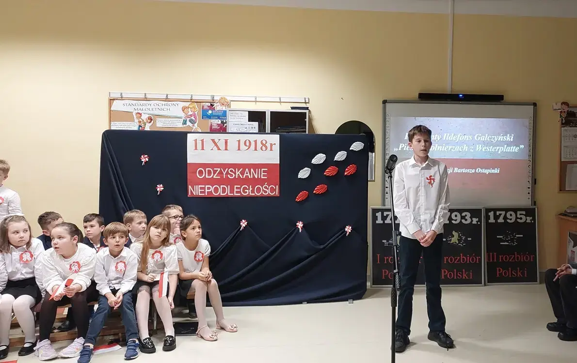  uczeń klasy szóstej recytuje wiersz a z jego prawej i lewej strony siedzą uczniowie klas młodszych i starszych biorący udział w wieczornicy