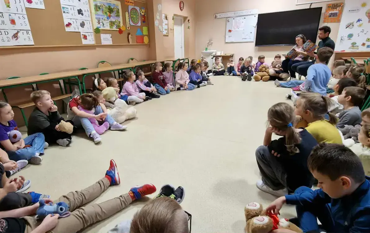 dzieci z klas pierwszych siedzą w kole na podłodze i słuchają treści książki czytanej przez autora i nauczycielkę