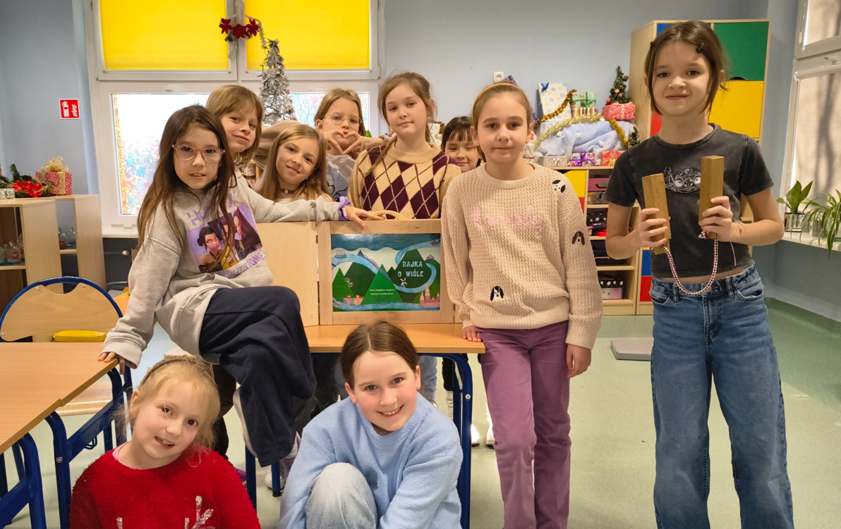Zdjęcie przedstawia uczennice klasy drugich i trzecich pozujące na tle teatrzyku kamishibai z Bajką o Wiśle.