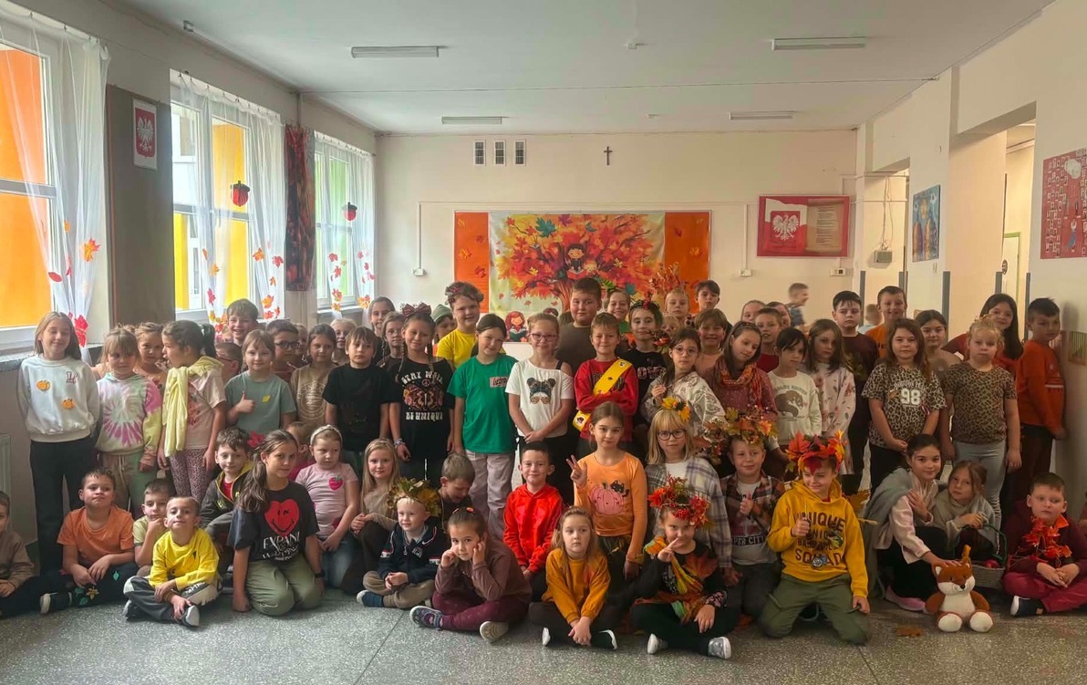 Grupa dzieci ubranych w jesienne stroje i ozdoby pozuje wspólnie na szkolnym korytarzu. W tle widoczne są jesienne dekoracje, kolorowe liście oraz prace plastyczne