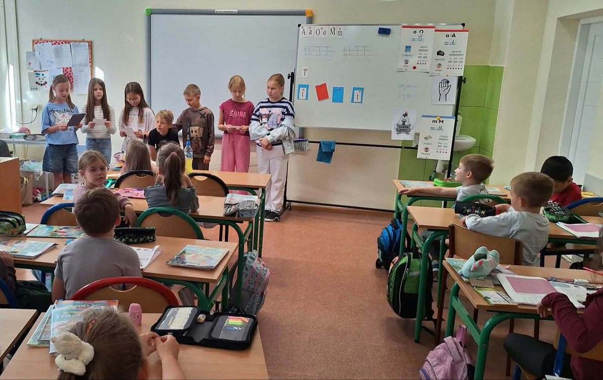 Uczniowie siedzą w klasie i słuchają kolegi czytającego książkę. Dzieci zgromadzone są w części sali, a na półkach w tle znajdują się książki i materiały edukacyjne.