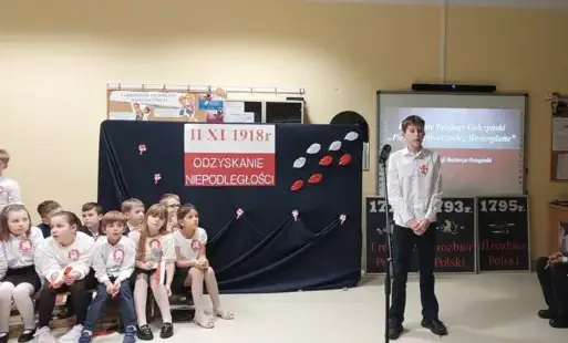  uczeń klasy szóstej recytuje wiersz a z jego prawej i lewej strony siedzą uczniowie klas młodszych i starszych biorący udział w wieczornicy