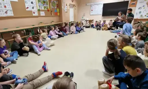 dzieci z klas pierwszych siedzą w kole na podłodze i słuchają treści książki czytanej przez autora i nauczycielkę