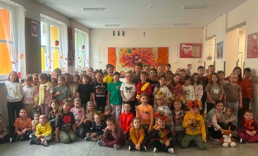 Grupa dzieci ubranych w jesienne stroje i ozdoby pozuje wspólnie na szkolnym korytarzu. W tle widoczne są jesienne dekoracje, kolorowe liście oraz prace plastyczne