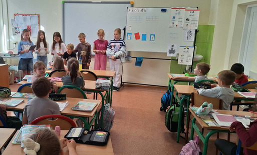 Uczniowie siedzą w klasie i słuchają kolegi czytającego książkę. Dzieci zgromadzone są w części sali, a na półkach w tle znajdują się książki i materiały edukacyjne.