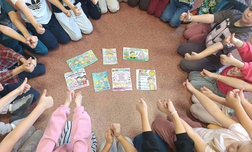 Uczniowie siedzą w kręgu na podłodze i pokazują kciuki uniesione w górę. W centrum kręgu znajdują się kolorowe materiały dotyczące przeciwdziałania przemocy rówieśniczej, między innymi plakaty z hasłami zachęcającymi do re